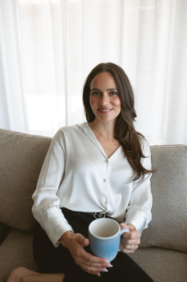 Daniella Simas Medeiros holding a ceramic mug, seated on a sofa in a softly-lit therapy room